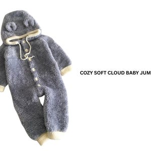 Könnte beinhalten: Ein weicher, grauer Baby-Overall mit Kapuze und kleinen Ohren. Der Kapuzen-Overall hat eine Knopfleiste vorne und cremefarbene Bündchen und Besätze. Der Text "COZY SOFT CLOUD BABY JUMPSUIT" wird angezeigt.