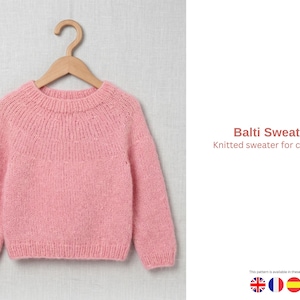 May include: A light pink knitted sweater for children, hanging on a wooden hanger. The sweater has a round neckline and long sleeves. The text "Balti Sweater Knitted sweater for children" is visible on the right side of the image.