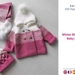 Puede incluir: Un conjunto de bebé tejido en rosa y blanco, que incluye una chaqueta con capucha, un gorro y una bufanda. La chaqueta tiene botones de madera y el gorro y la bufanda tienen pompones esponjosos. El conjunto está diseñado para el invierno.