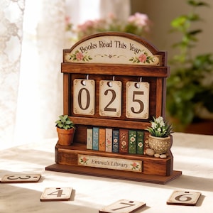 May include: Wooden bookshelf-style counter with the text "Books Read This Year" and "Emma's Library". The counter displays "025" and includes miniature books and potted plants. Additional number tiles are scattered around.
