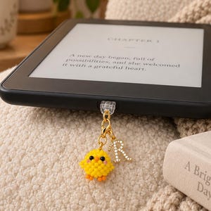 May include: A black e-reader device with a screen displaying text starting with Chapter 1. A gold-toned charm is attached to the bottom edge, featuring a small yellow beaded chick figure and a letter R charm encrusted with clear crystals.