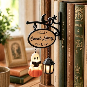Op de afbeelding: Een zwart metalen wandbord met een crèmekleurig ovaal plaatje met de tekst "Emma's Library", een boekicoon, een maansikkel en sterren. Een kleine geest die op een pompoen leest en een zwarte lantaarn hangen aan het bordje.