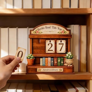 以下が含まれることがあります： 「Books Read This Year」の文字と日付「27」が表示された、本棚スタイルの木製カレンダー。 ミニチュアの本と小さな鉢植えの植物が置かれた棚があります。「Sarah's Library」の文字も表示されています。