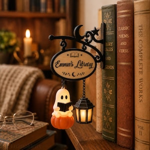 May include: A black metal wall-mounted sign bracket with a moon and star design, featuring an oval wooden plaque inscribed with "Emma's Library" and an open book icon. A small glowing ghost sitting on a pumpkin and a lantern hang from the sign.