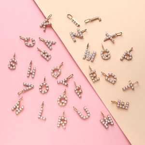 Peut inclure: Une collection de breloques alphabet dorées, chacune ornée de petites perles blanches. Les lettres sont disposées sur un fond rose et beige. Chaque breloque est munie d'une petite boucle pour l'attacher à un collier ou un bracelet.