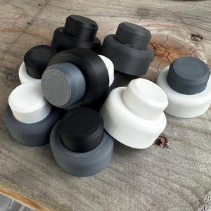 May include: A collection of mushroom-shaped bottle stoppers in various colors, including black, white, and gray. The stoppers are made of a solid material and have a textured top. They are arranged on a wooden surface.