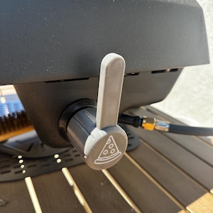 May include: Close-up of a gray pizza oven control knob with a white pizza slice graphic. The knob is attached to a black oven, with a gray handle. The oven is on a wooden table.