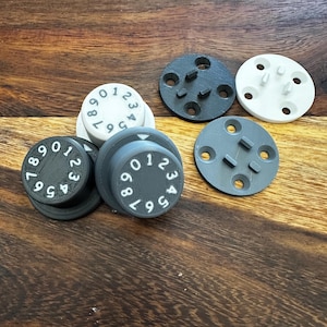 Peut inclure: Cinq cadrans de serrure à combinaison ronds en plastique noir et blanc avec les chiffres de 0 à 9. Les cadrans sont sur une surface en bois.