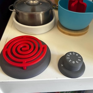 May include: A toy stovetop with a black and gray burner with a red spiral design. The stovetop has a black knob with white numbers 1 through 8.