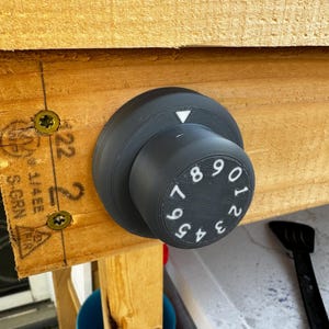 May include: A black plastic combination lock with white numbers 0-9. The lock is mounted on a wooden surface.