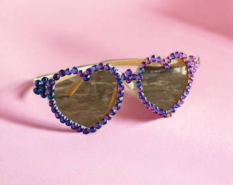 May include: Heart-shaped sunglasses with a gold frame and purple and blue rhinestones.