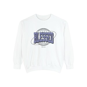 Op de afbeelding: Witte sweatshirt met het woord "BLESSED" in een paarse, retro lettertype, tegen een wereldbol ontwerp. De tekst "John 10:29" staat eronder. De sweatshirt heeft een ronde hals en lange mouwen.