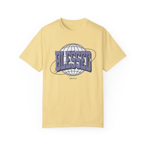 Op de afbeelding: Lichtgele t-shirt met het woord "BLESSED" in een paars en wit ontwerp, tegen een witte wereldbolafbeelding. De tekst "John 3:29" staat eronder. Een casual kledingstuk.