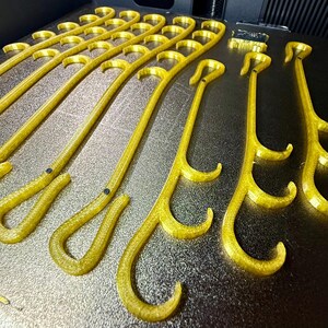 May include: A collection of translucent yellow, 3D-printed plastic hooks arranged in rows. Each hook features a long, curved handle with multiple smaller, integrated hook protrusions along the side and a small black circular inlay near the base.