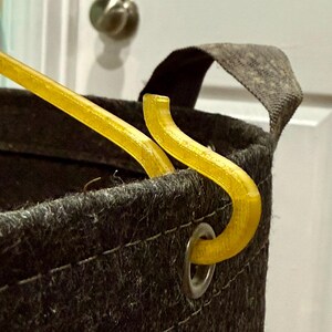 May include: A dark gray felt storage basket featuring a silver metal grommet on the upper rim. A translucent yellow plastic hook is inserted through the grommet, with a matching yellow plastic rod extending diagonally from the basket opening.