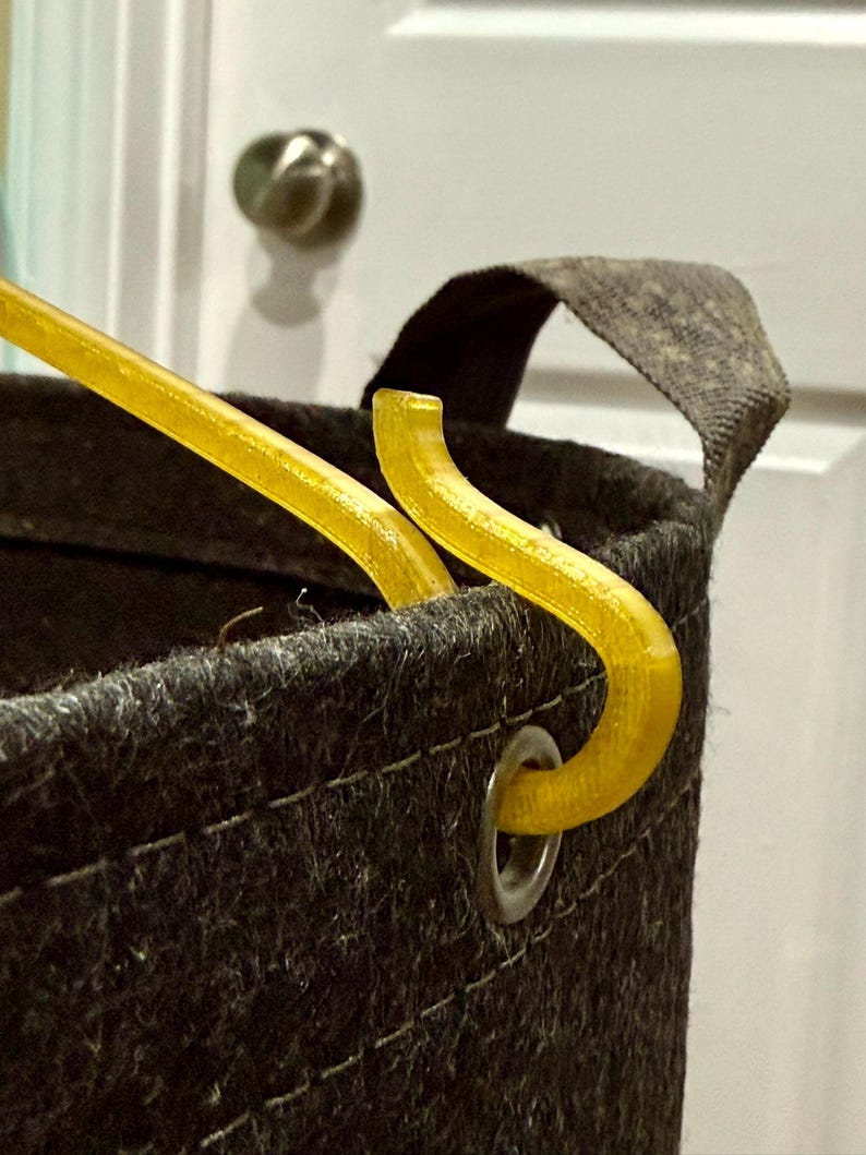 May include: A dark gray felt storage basket featuring a silver metal grommet on the upper rim. A translucent yellow plastic hook is inserted through the grommet, with a matching yellow plastic rod extending diagonally from the basket opening.