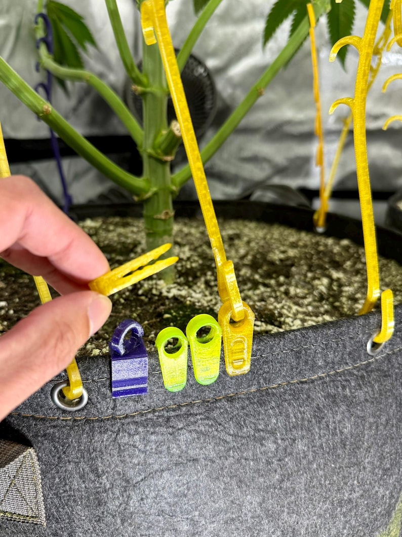 May include: A set of yellow and purple plastic plant training clips and hooks attached to the rim of a grey fabric grow bag. The clips feature a small loop design for securing plant stems, with one yellow clip held between fingers.