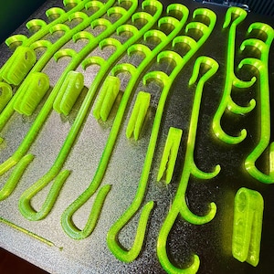 May include: A collection of neon green 3D printed plastic hooks and clips arranged on a dark textured build plate. The items feature curved hook ends and rectangular clip bases with visible layer lines from the additive manufacturing process.