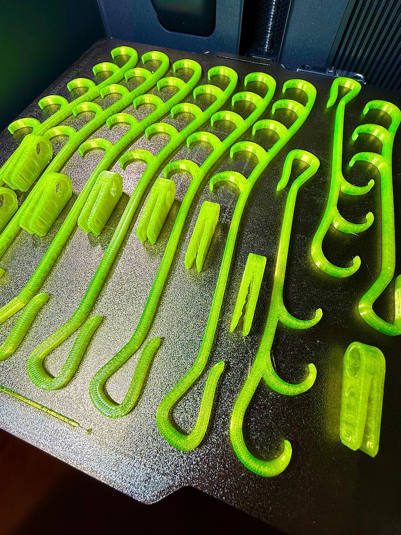 May include: A collection of neon green 3D printed plastic hooks and clips arranged on a dark textured build plate. The items feature curved hook ends and rectangular clip bases with visible layer lines from the additive manufacturing process.