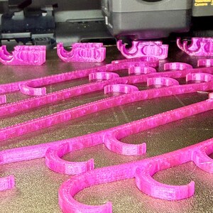 May include: A 3D printer is actively creating multiple magenta plastic objects on a print bed. Each object features a long, straight bar with several evenly spaced, curved hook-like protrusions extending from one side, showing a layered texture.