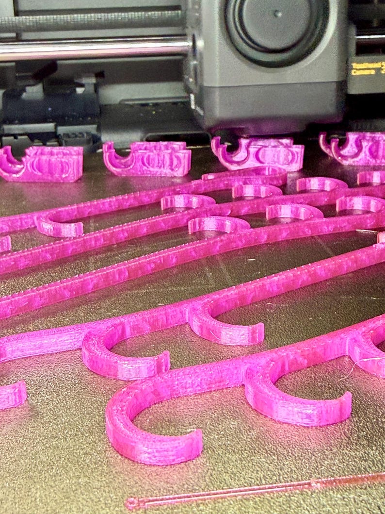 May include: A 3D printer is actively creating multiple magenta plastic objects on a print bed. Each object features a long, straight bar with several evenly spaced, curved hook-like protrusions extending from one side, showing a layered texture.