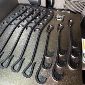 May include: A collection of black, multi-hook hangers arranged on a flat surface. Each hanger features a curved top hook and multiple lower hooks along its length, with small metallic fasteners visible at the connection points of the hooks.