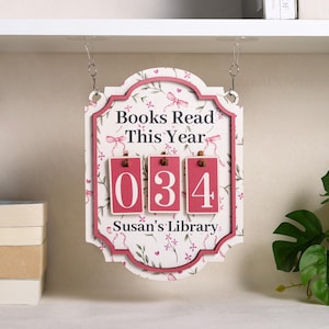 May include: A decorative wooden sign with a floral pattern and the text "Books Read This Year." It features a counter displaying "034" and the text "Susan's Library." The sign is framed in pink and hangs from metal chains.