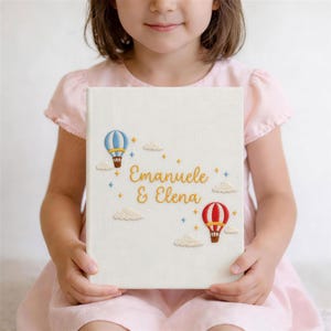 May include: A cream-coloured book featuring embroidered hot air balloons in blue and red, clouds, and the names "Emanuele & Elena" in gold lettering. The book is held by a person wearing a pink dress.