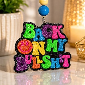May include: A hanging car air freshener featuring a textured, glittery black background with multi-colored, raised lettering that reads BACK ON MY BULLSHIT, with a pink smiley face replacing the O. It hangs from a pink string with three beads.