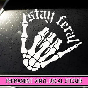 May include: White vinyl decal sticker with a skeleton hand giving the shaka sign and the text "Stay Weird" in a gothic font.