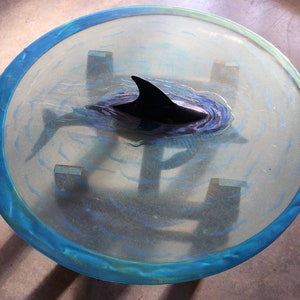 Puede incluir: Una mesa redonda de cristal con un borde azul. La mesa tiene una escultura de delfín negro con un diseño de agua azul y blanco.