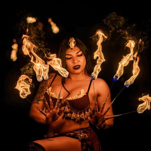 May include: A woman wearing a brown and gold costume with fringe is performing a fire dancing act. She is holding multiple flaming sticks in each hand, creating a circle of fire around her.