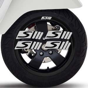 Può includere: Primo piano di una ruota di moto nera con pneumatico. La ruota presenta un cerchio nero e una serie di decalcomanie bianche e grigie con le lettere "S" e "III". La moto è bianca.