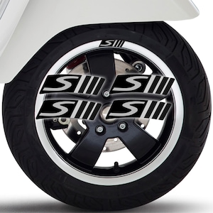 Può includere: Primo piano di una ruota di scooter con pneumatico e cerchio neri. La ruota presenta decalcomanie nere e argentate con la lettera "S" e tre linee verticali. Lo scooter è bianco.