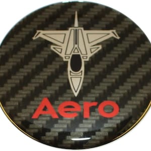 Könnte beinhalten: Ein runder Aufkleber aus schwarzem und grauem Carbon mit einer weißen Silhouette eines Kampfjets und dem Wort "Aero" in roten Buchstaben.