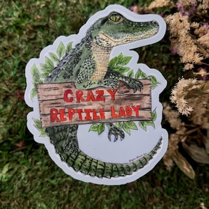 Könnte beinhalten: Aufkleber mit einer detaillierten Illustration eines grünen Alligators, der ein Holzschild mit dem roten Text "CRAZY REPTILE LADY" hält. Der Alligator ist von grünen Blättern umgeben, mit einem weißen Rand.