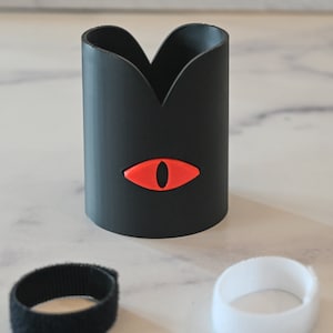 Op de afbeelding: Een zwarte 3D-geprinte beker met een rood oogontwerp. De beker wordt geleverd met twee zwarte en witte klittenbandstrips.