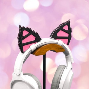 May include: White headphones with a pink and black cat ear headband on a black stand. The headphones are on a stand against a pink and white bokeh background.