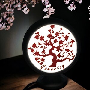 Op de afbeelding: Een ronde, zwarte lamp met een kersenbloesemboomontwerp in rood. De boom is gecentreerd op een witte achtergrond, met het woord "Gamertag" eronder. De lamp staat op een zwarte voet en een houten oppervlak.