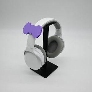 May include: White wireless headphones with a purple bow on the headband, sitting on a black stand.