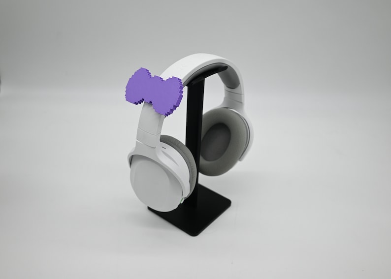 May include: White wireless headphones with a purple bow on the headband, sitting on a black stand.