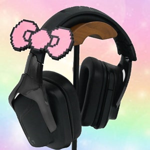 May include: Black gaming headphones with a pink pixelated bow on the headband. The headphones are on a wooden stand.