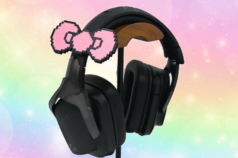 May include: Black gaming headphones with a pink pixelated bow on the headband. The headphones are on a wooden stand.