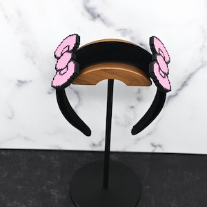 May include: Black headband with two pink pixelated bows on either side. The headband is on a wooden stand.