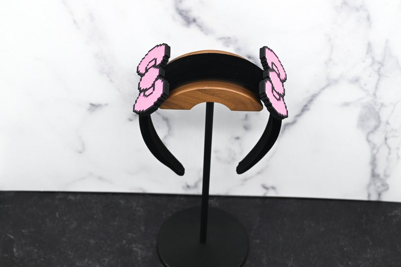 May include: Black headband with two pink pixelated bows on either side. The headband is on a wooden stand.