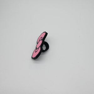 May include: A pink and black ring with a three-dimensional design. The ring is made of plastic and has a smooth finish.