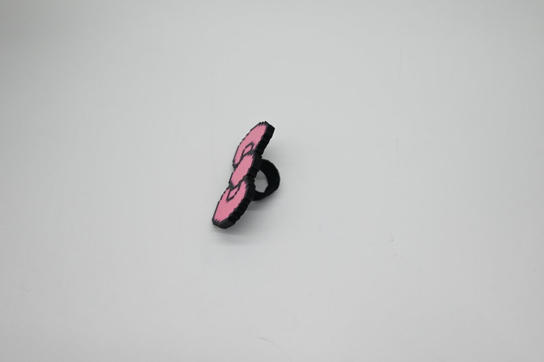 May include: A pink and black ring with a three-dimensional design. The ring is made of plastic and has a smooth finish.