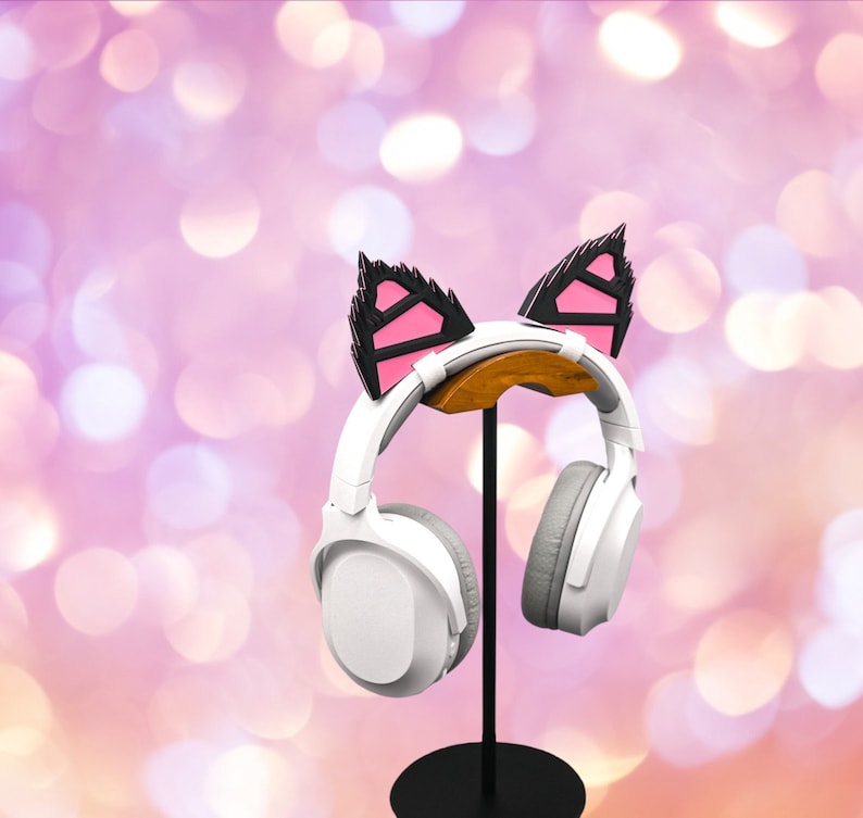 May include: White headphones with a pink and black cat ear headband on a black stand. The headphones are on a stand against a pink and white bokeh background.