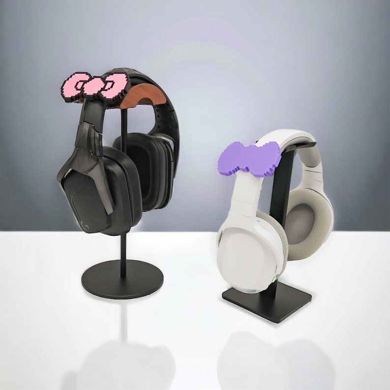 May include: Two black headphone stands with different colored bow-shaped accessories. The stand on the left has a pink bow and the stand on the right has a purple bow.