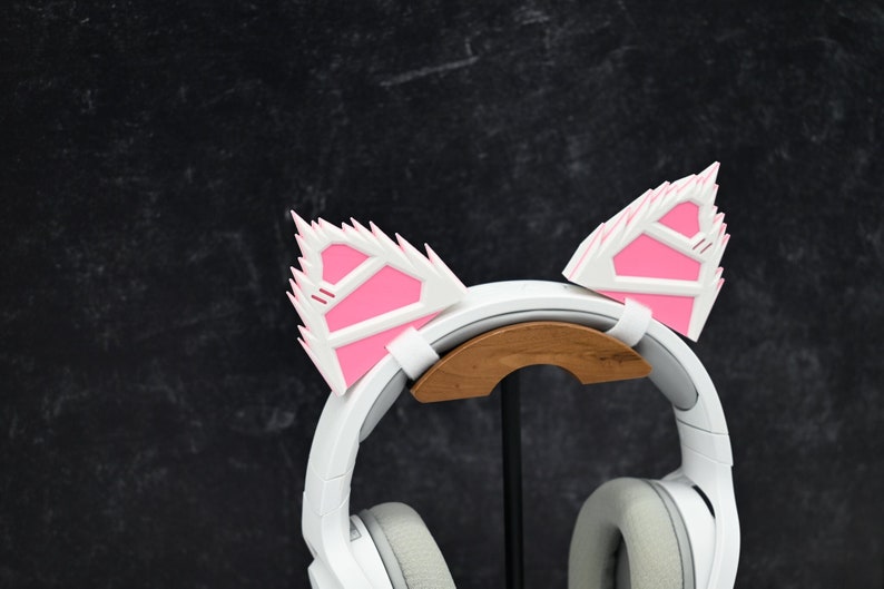 May include: White headphones with pink cat ears attached to the headband. The headphones are on a wooden stand.
